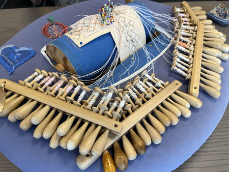 Närbild av en spetsknypplingsuppsättning med trådar, pinnar och mönster på en blå kudde.