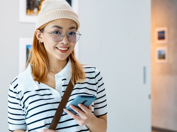 Ung kvinna med glasögon och mössa står leende i ett konstgalleri och håller en mobiltelefon.