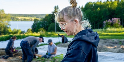 En kvinna med glasögon arbetar på en odlingsmark med flera personer i bakgrunden, omgiven av grönska.