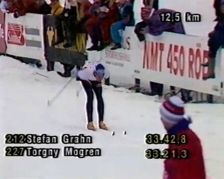 Skidåkare tävlar i längdskidåkning, omgiven av publik och snö, med resultat visas på skärmen.