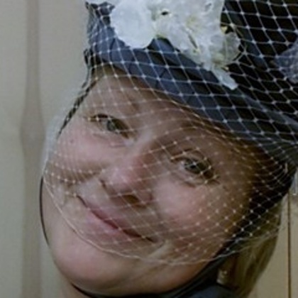 Person med ljusblå kavaj och vit blus bär en hatt med vita blommor och nätdekoration.