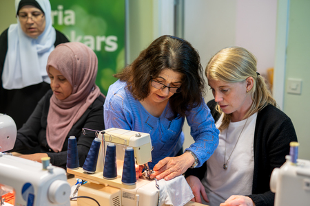 Fyra kvinnor sitter vid symaskiner och arbetar tillsammans i ett ljust rum.