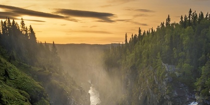 Högt vattenfall i en djup grön dal omgiven av skog vid solnedgången.