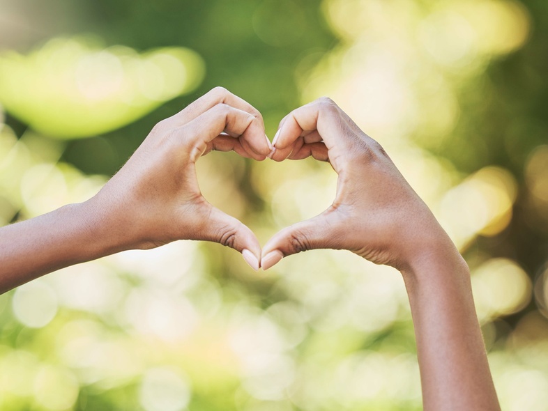 Två händer formar ett hjärta mot en suddig, grön bakgrund utomhus.