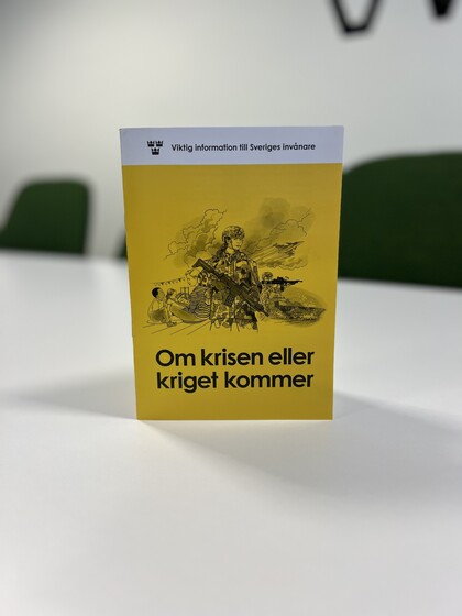 En gul broschyr med texten 'Om krisen eller kriget kommer' står på ett vitt bord i ett mötesrum.