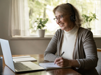 Äldre kvinna med hörlurar deltar i videosamtal vid laptop, antecknar i block vid ett bord.