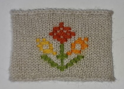 Stickad lapp med broderat blommönster i rött, gult och grönt på beige bakgrund.