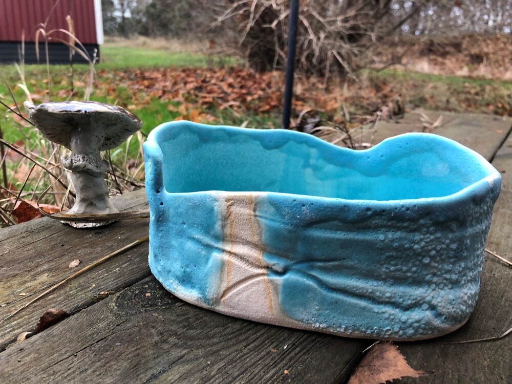 En blå keramikskål och en liten svampformad skulptur står på ett träbord utomhus med höstlöv runtom.