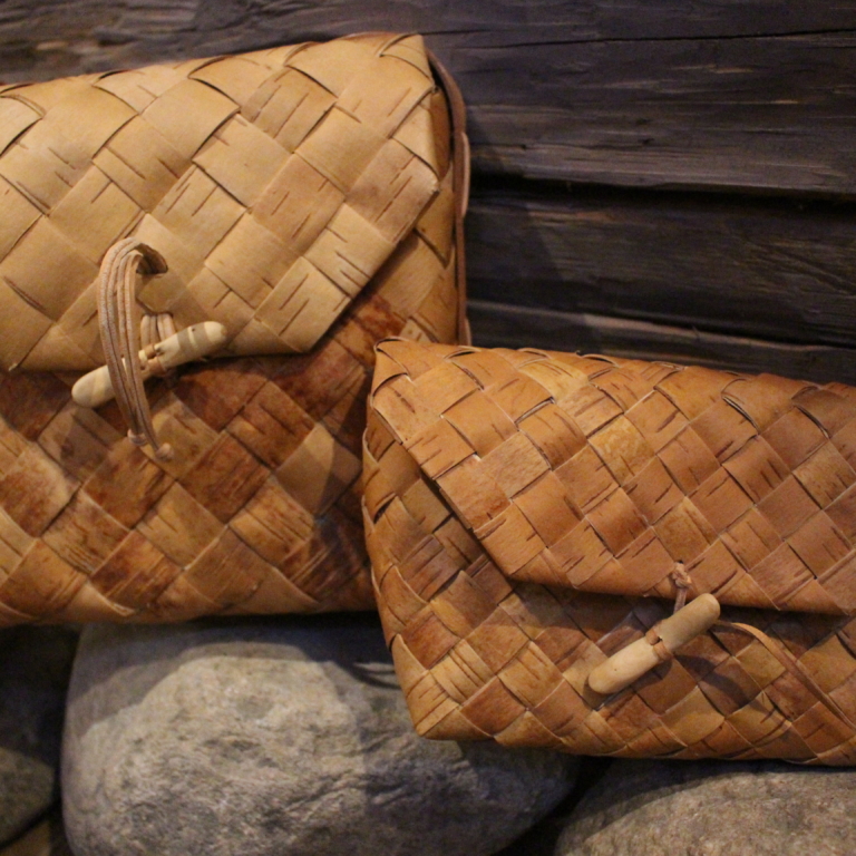 Två handgjorda väskor av flätad näver ligger på stenar framför en trävägg.