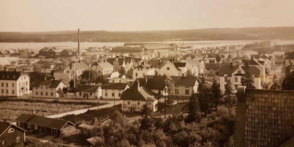 Gammalt svartvitt sepiafärgat foto över en småstad med trähus, fabriker och vatten i bakgrunden.