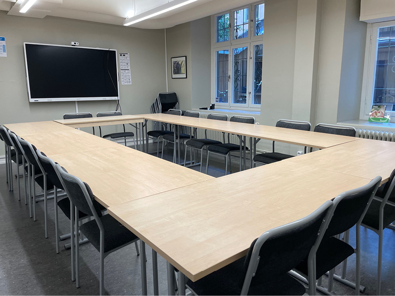 Ett tomt konferensrum med U-formade bord, svarta stolar och en stor svart tavla på väggen.