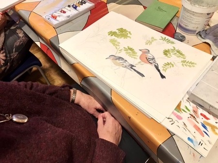 En person målar fåglar och blad med akvarellfärger vid ett bord täckt med färgglad duk.
