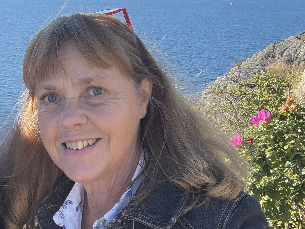 Person med långt hår står utomhus vid vatten och klippor, blommor syns i bakgrunden.