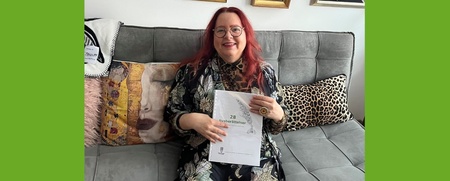 Kvinna med rött hår och glasögon sitter i soffa och håller i ett papper, omgiven av färgglada kuddar.