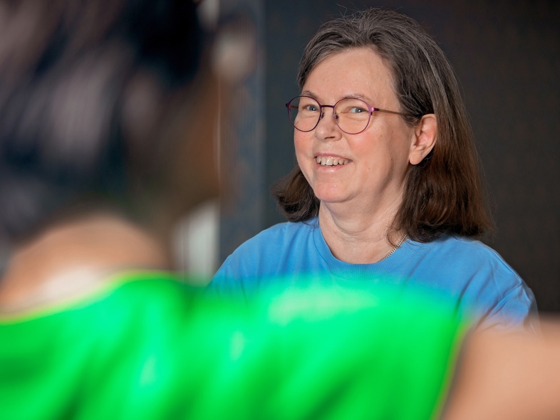 Leende kvinna med glasögon och blå tröja sitter inomhus, suddig person i förgrunden.