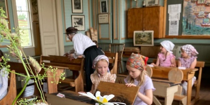 Barn i gammaldags kläder sitter vid bänkar i ett historiskt klassrum med blommor i förgrunden.