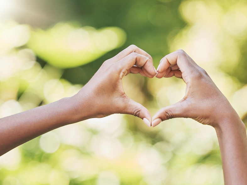 Två händer formar ett hjärta mot en suddig, grön bakgrund utomhus.