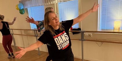Kvinna dansar glatt i en dansstudio med trägolv och speglar, iförd svart t-shirt med texten 'DANS STUDIO'.