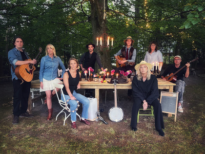 En grupp människor sitter och står runt ett bord utomhus, omgivna av träd och levande ljus.