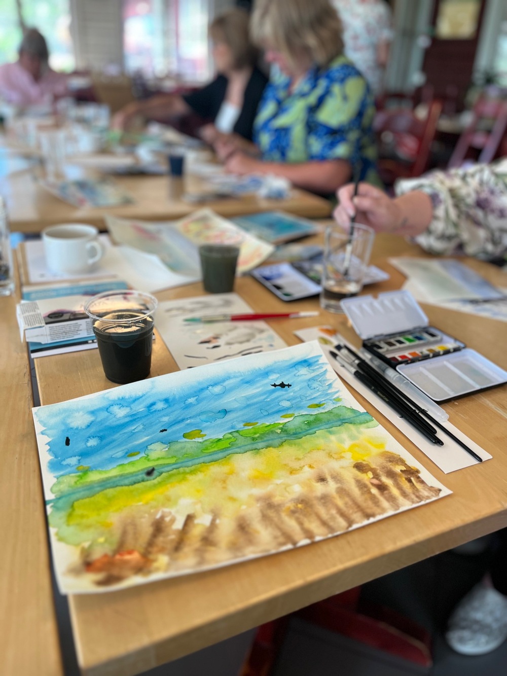Personer målar akvarell vid ett bord med konstmaterial och färgglada målningar.