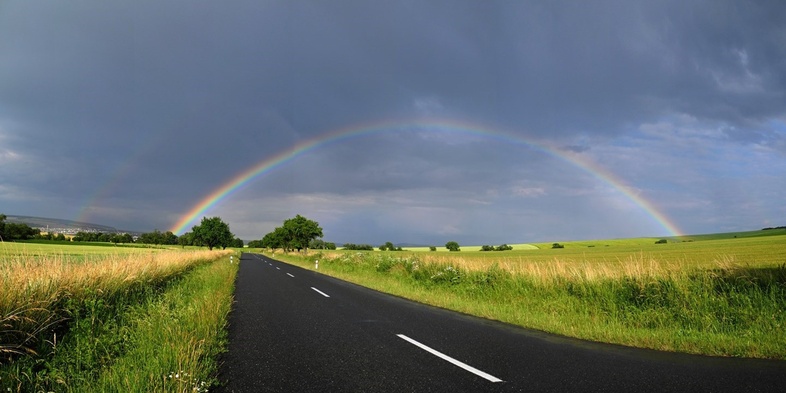 En landsväg leder genom gröna fält under en mörk himmel med en tydlig regnbåge.