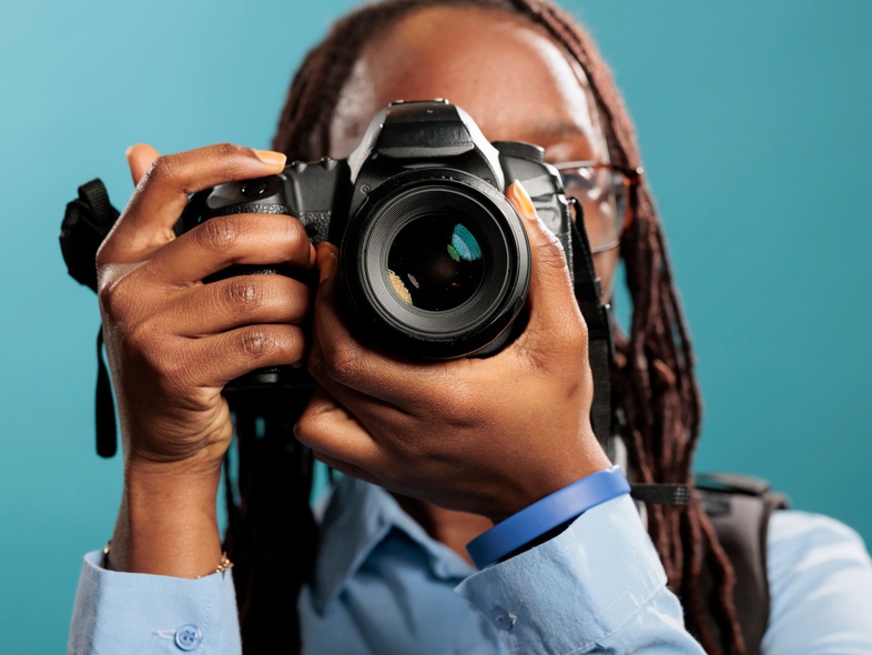Person med glasögon och blå skjorta håller en kamera framför ansiktet mot en blå bakgrund.