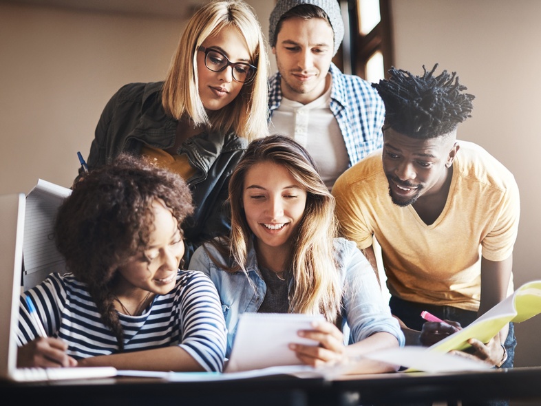 Fem unga vuxna studerar tillsammans vid ett bord, tittar på papper och ler.