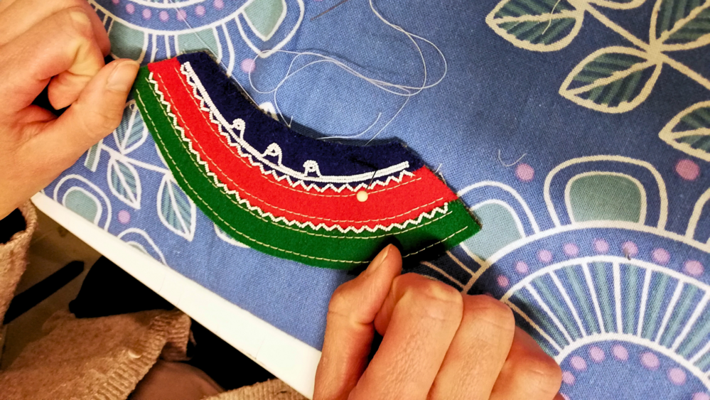 Två händer syr färgglatt tyg med broderi på ett blått mönstrat underlag.