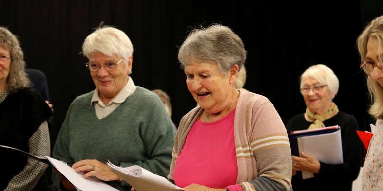 Fem äldre kvinnor står och sjunger tillsammans i ett rum, håller papper och ser glada ut.