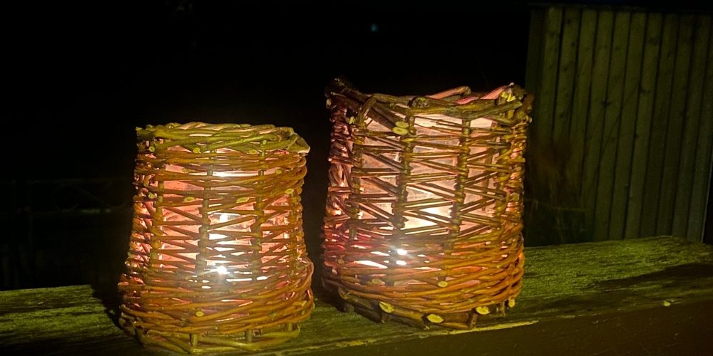 Två tända ljuslyktor av flätad korg står på ett träbord utomhus i mörkret.