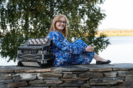 Person i blå klänning sitter på en stenmur vid vatten, bredvid ett dragspel med texten 'Giulietti'.