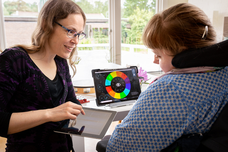 Två personer använder surfplattor och en färgglad kommunikationsapp vid ett bord inomhus.