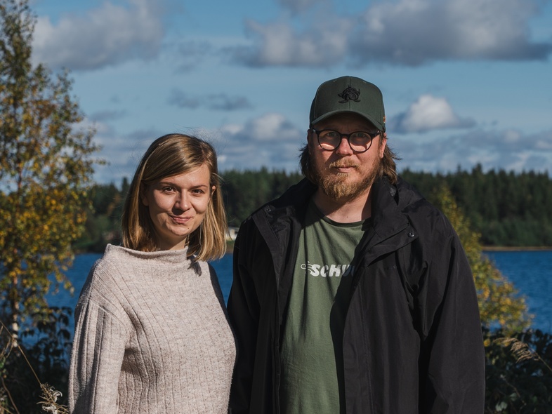 En kvinna och en man står utomhus vid en sjö med skog i bakgrunden under en solig dag.