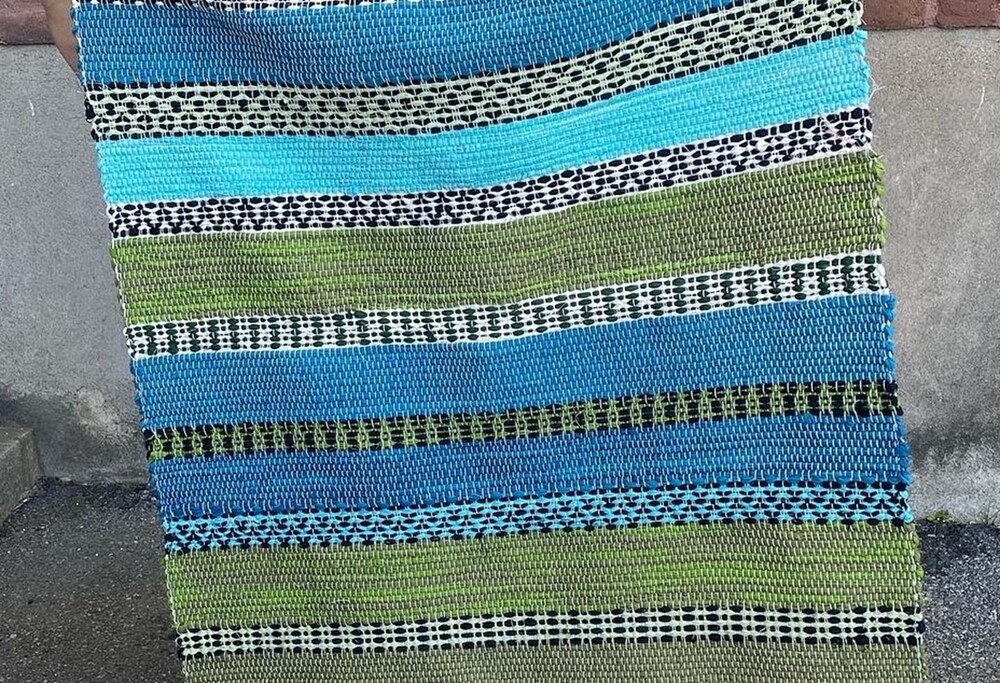 En handvävd matta med ränder i blått, grönt, svart och vitt står lutad mot en vägg utomhus.