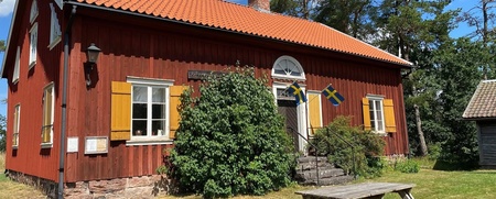 Röd trävilla med gula fönsterluckor, svenska flaggor och picknickbord i trädgård, solig dag.