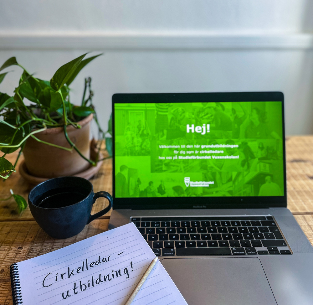 En laptop med grön välkomstskärm, anteckningsblock, penna, kaffekopp och krukväxt på ett träbord.