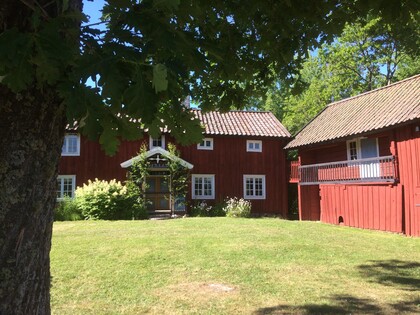 Röd trähusgård med vita fönster, grön gräsmatta och träd i förgrunden en solig dag.