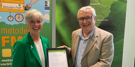 Ulrika Heie och Björn Körlof håller ett diplom tillsammans vid en monter med gröna och färgglada affischer.