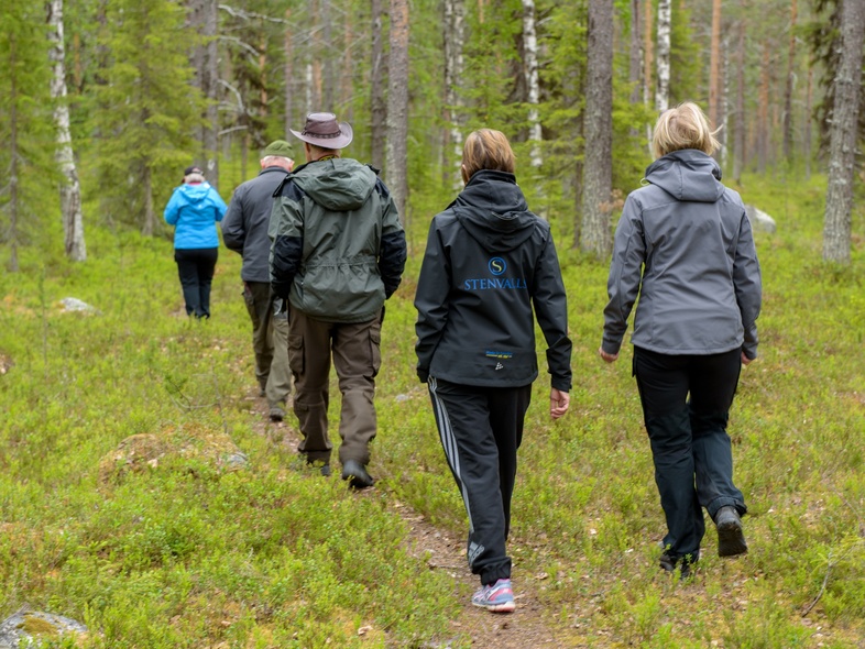 En grupp människor vandrar på en stig genom en grön skog, sedda bakifrån.