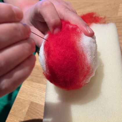 En person nålfiltar röd ull på en vit boll ovanpå en skumgummibit på ett träbord.