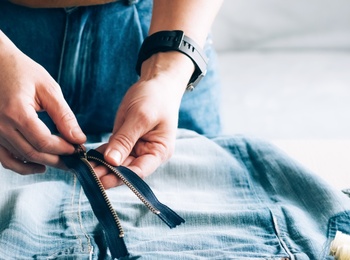 Person arbetar med att sy om ett par jeans, med dragkedja, måttband och sytråd på bordet.