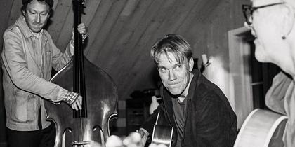 Tre personer spelar akustiska instrument i ett rum med träpanel, omslag till albumet 'Medan Viskan rinner' av Claes Pihl.