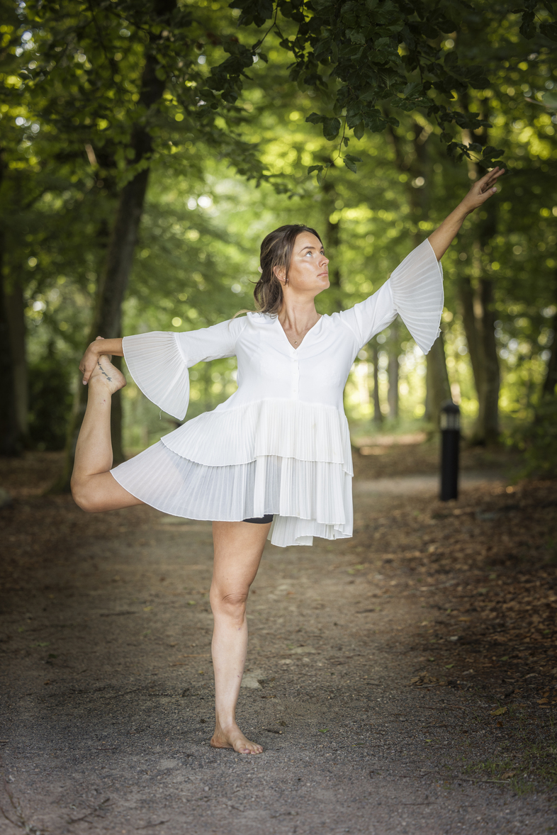 Kvinna i vit klänning gör en yogaposition på en skogsstig, omgiven av gröna träd.