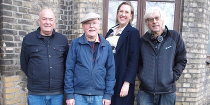 Fyra personer utan instrument står utomhus framför en tegelvägg, tre män och en kvinna.