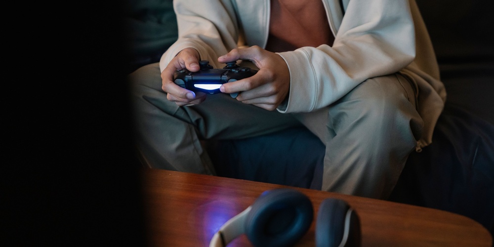 Person sitter på soffa och spelar tv-spel med en handkontroll, hörlurar ligger på bordet framför.