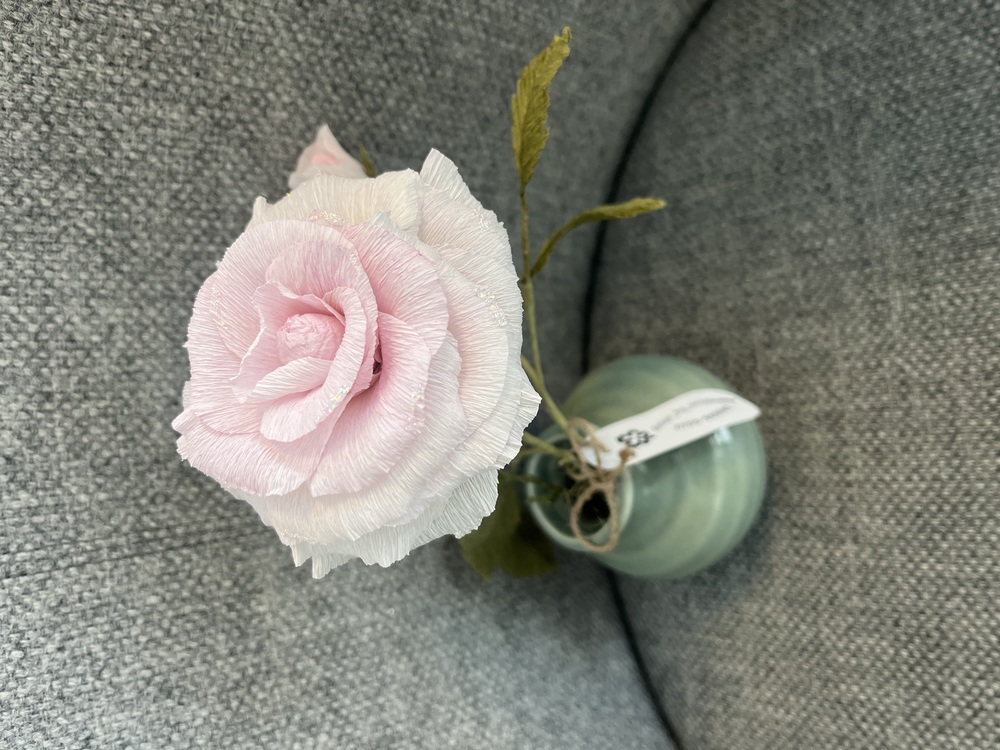 En ljusrosa konstgjord ros i en liten grön vas, placerad på en grå tygsoffa.
