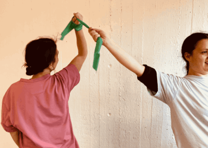 Två personer håller i ett grönt band och sträcker upp armarna mot varandra framför en ljus vägg.