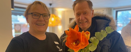 Två glada personer står inomhus, en håller en stor orange blomma.