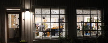 Ett upplyst rum syns genom stora fönster på ett hus i mörkret, med människor och konstnärsmaterial inuti.