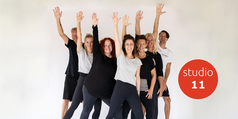 En grupp glada vuxna i träningskläder poserar med armarna uppsträckta framför en vit bakgrund.